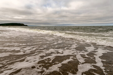 Gentle ocean waves foam across the sandy shoreline beneath a moody sky, with distant headland visible on the horizon, dramatic atmosphere. Sea of Okhotsk. Sakhalin Island