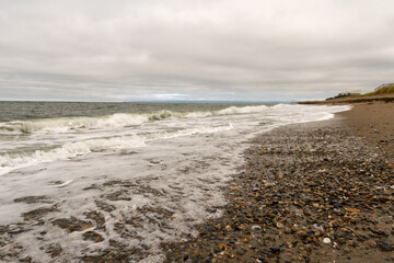 Waves roll onto shoreline scattered with wet pebbles and stones beneath cloudy sky, creating natural seascape that combines ocean movement with textured beach details. Sea of Okhotsk Sakhalin Island