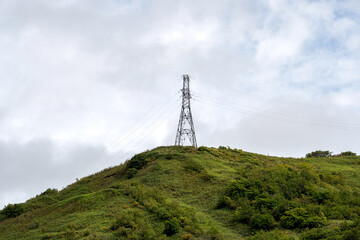 A single electricity transmission tower rises on a grassy hillside, its steel structure contrasting with the lush green vegetation and cloudy overcast sky
