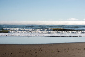 Rolling waves crash against a sandy beach while a low fog bank stretches across the horizon, separating the sea from the clear morning sky. Sea of Okhotsk, Sakhalin Island