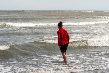 Girl wearing a bright red jacket wades into the shallow sea waves, facing the horizon while enjoying the refreshing coastal water. Tikhaya Bay in the Sea of Okhotsk, Sakhalin Island