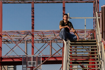 Man sits in control on stair rail within a red metal structure, gazing confidently toward camera while demonstrating balance and spatial awareness. Precision in urban acrobatic movement. Parkour 