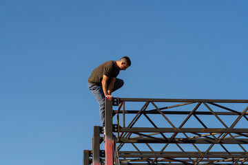 Male freerunner crouches carefully on narrow edge of tall red steel training frame, demonstrating precision, strength, and focus against blue sky. Precision in urban acrobatic movement. Parkour perfor