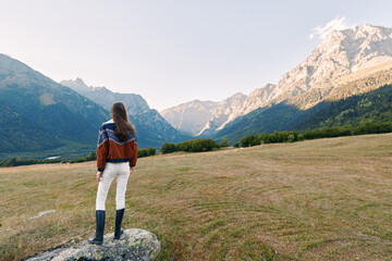 Woman in a patterned sweater stands on a rock in a wide meadow facing distant mountains, hiking across a peaceful landscape with boots, valley view and serene natural scenery. © SHOTPRIME STUDIO