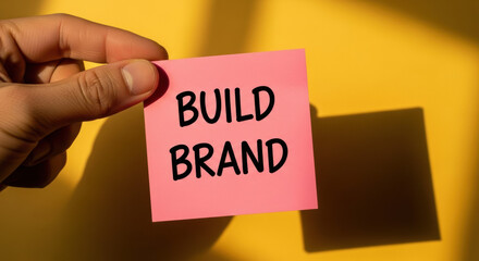 Hand Holding Pink Sticky Note With Build Brand Text Against Yellow Background With Dramatic Shadows