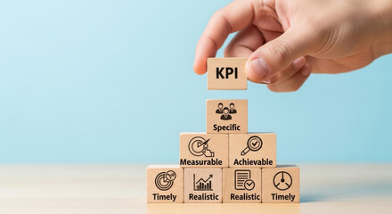 A hand carefully places a wooden block labeled KPI on top of a pyramid of blocks with business icons. The blocks symbolize business strategy and growth.