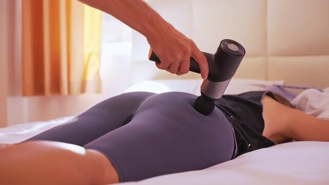 Woman lying on bed as massage gun presses on Hip in tight leggings with a medium low angle highlighting visible slow motion pulsing, Deep Tissue Concept
