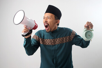 Angry Asian Muslim man in a green traditional outfit shouting into a megaphone while holding an alarm clock in his hands. Isolated on white background