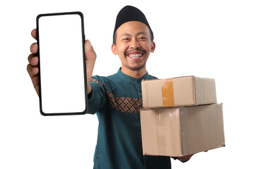 Asian Muslim man carrying a stack of cardboard boxes while showing copy space on his phone screen with a blank white display, ideal for delivery, online shopping concept, isolated on white background