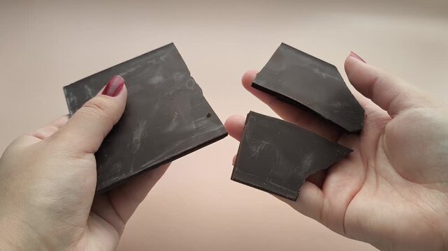 Broken pieces of dark chocolate from cocoa beans in a woman's hand on a pink background.
