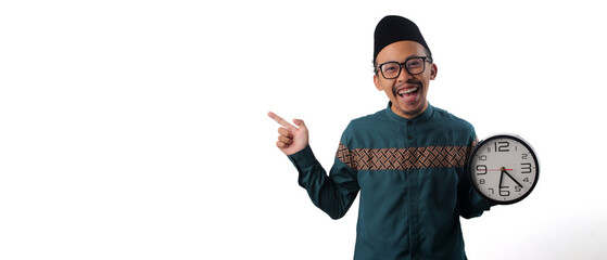 Excited Asian Muslim man wearing a green traditional outfit, pointing at empty copy space while holding a clock in his other hand. Isolated on a white background. Time announcement, countdown concept