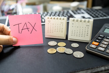 Hand holding TAX reminder note with coins and 2026 calendar for tax season and financial planning concept. tax season, financial burden, calculation of tax returns, and economic challenges.