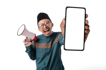 Asian Muslim man in a green traditional outfit showing a smartphone with an empty white screen with copy space to the camera while holding a megaphone in the other hand. Isolated on white background.