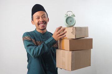 Happy Asian Muslim man wearing a green traditional outfit and black songkok, smiling at the camera while holding a stack of cardboard boxes with Alarm Clock on Top. Isolated on a white background