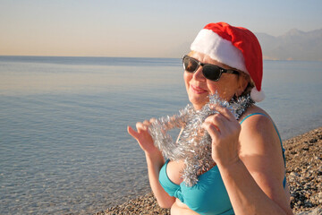 Funny senior woman in santa claus hats and sunglasses wearing in swimsuit on the sea beach, women dancing and wishes a merry Christmas and happy new year
