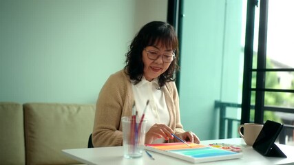 Smiling pretty senior asian woman painting and coloring on a canvas at home with creative lifestyle and healthy aging with mental wellness for retirement, elderly female enjoy leisure activity.
