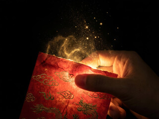 Macro shot of hand holding magical Chinese red envelope with rising golden dust and glowing sparkles against black background.