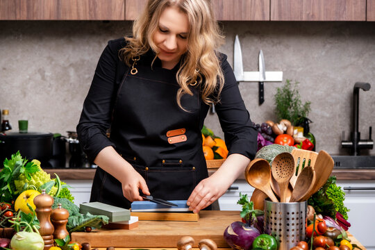 Focused blonde woman in black chef apron sharpens kitchen knife on whetstone at wooden table with vegetables in bright modern kitchen. Cooking, culinary skills, healthy lifestyle concept.