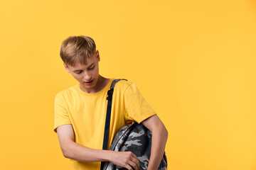 Student wearing a yellow t shirt with a camouflage backpack against a vibrant yellow background,...