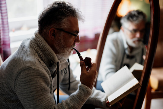 Man reads book while smoking pipe in cozy room with mirror reflection