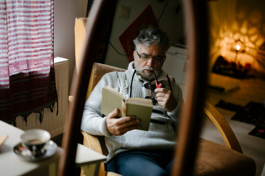 Man sitting in chair reading a book while enjoying a pipe