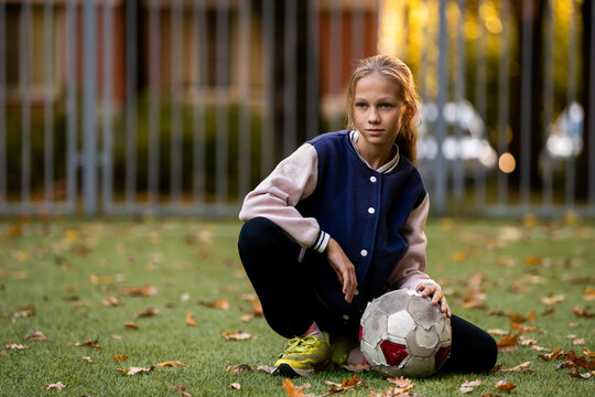 girl squats with a battered soccer ball on a green field covered