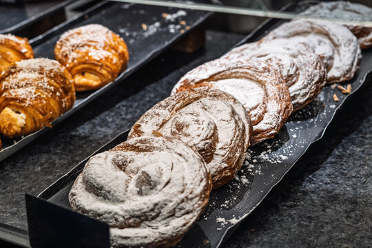 Close-up of sweet spiral pastries or ensaimadas dusted with powdered sugar arranged on a black tray in a bakery counter.