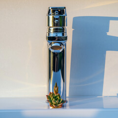 A sleek silver vaporizer with a small succulent plant at its base, standing upright on a white surface, with a shadow cast beside it