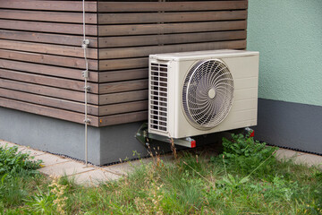 Air conditioning unit mounted on exterior wall of modern building, surrounded by green grass and wooden paneling, showcasing outdoor cooling technology and design aesthetics