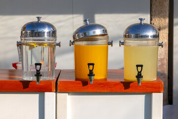 Three large glass beverage dispensers with chrome lids and taps on a wooden counter, filled with water, orange juice and lemonade, ready for self service at a cafe or event
