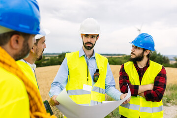 Team of engineers discussing blueprints and project plans at a wind farm construction site, collaborating on sustainable development. Renewable energy and industrial workforce concept