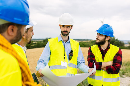 Team of engineers discussing blueprints and project plans at a wind farm construction site, collaborating on sustainable development. Renewable energy and industrial workforce concept - Powered by Adobe