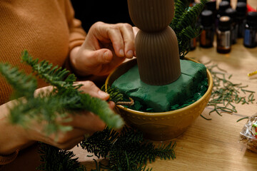 Close-up of woman's hands creating a Christmas floral arrangement using green floral foam, fresh fir branches, and essential oils on a wooden table.
