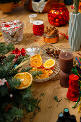 Festive Christmas table setting with dried orange slices in a glass bowl, cinnamon sticks, red ornaments, and decorative candles on a wooden background.