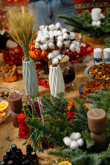 Festive Christmas workshop table with cotton flowers, wheat stalks in vases, candles, fir branches, and essential oil bottles prepared for holiday decorating.