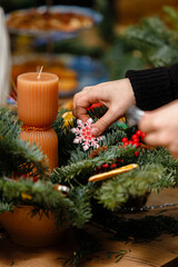 Close-up of a person's hands adding a white and red wooden snowflake ornament to a handmade Christmas centerpiece with fir branches and candles.