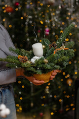 A person holding a beautiful handmade Christmas floral arrangement in a ceramic pot featuring a white candle, pine branches, cotton, and cones. holiday decor with blurred lights in the background.