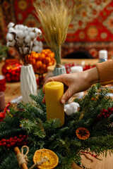 Close-up of a hand placing a yellow honeycomb beeswax candle into a lush Christmas arrangement. Artisanal holiday floristry workshop featuring natural materials like cotton flowers and dried oranges.