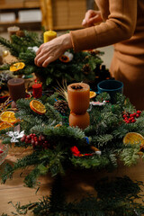 Close-up of a woman in a cozy beige sweater assembling a handmade Christmas wreath with a candle, pine cones, and dried oranges. Professional florist creating festive holiday home decor.
