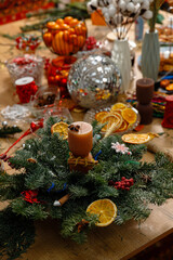 Handmade Christmas centerpiece with pine branches, dried orange slices, and a stylish candle on a festive wooden table. Cozy holiday DIY home decor concept with a blurred festive background.