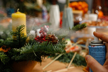 Close-up of a hand spraying artificial snow from a can onto a handmade Christmas floral arrangement. Creative process of making DIY holiday decorations with pine cones and berries.