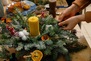 Close-up of a person's hands creating a festive Christmas centerpiece with evergreen branches, a yellow beeswax candle, cotton bolls, dried orange slices, and red berries on a wooden table.