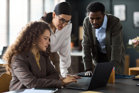 Young adult Caucasian woman sitting at desk using laptop while young Caucasian woman and Black man standing beside her, observing screen and discussing project