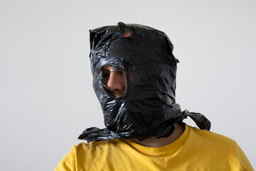 Portrait of an adult man in a yellow shirt with a black plastic bag over his head, cut for breathing and vision. Concept of hostage situation and abduction threat on a minimal light background