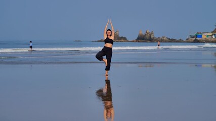 Full-length shot of female practicing yoga on wet sand with a strong grounded stance. The coastal background and open sky provide clean copy space for wellness and travel concepts.