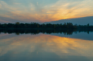 Sunset over a calm lake, reflection and trees in the background in the early evening.