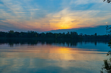 Sunset over a calm lake, reflection and trees in the background in the early evening.