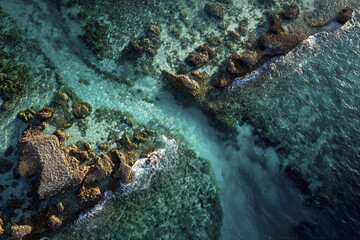 Aerial view of ocean water with coral reef formations and rocky structures creating natural patterns under sunlight, showcasing vibrant blue and green shades with gentle waves