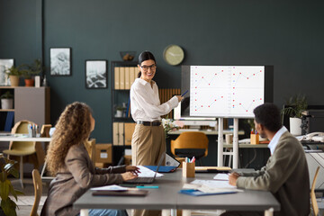 Caucasian woman standing and presenting data on digital screen to diverse young adult colleagues sitting at table in modern office, engaging in business meeting, discussing charts
