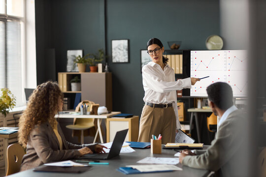 Caucasian young woman presenting data on screen to diverse colleagues in modern office, gesturing toward chart while explaining business statistics during meeting